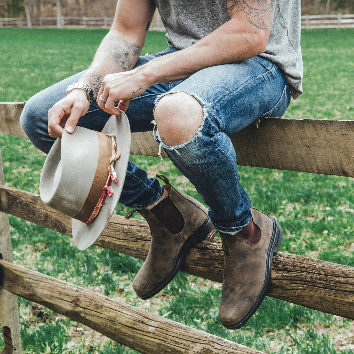 Person sitting on a wooden fence wearing ripped jeans and brown 1306 Blundstone boots, holding a hat.