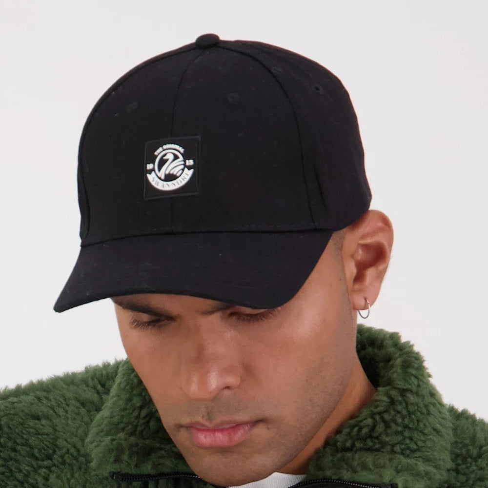 A person wearing a black Swanndri Swanni baseball cap with an adjustable back buckle and a white logo on the front.