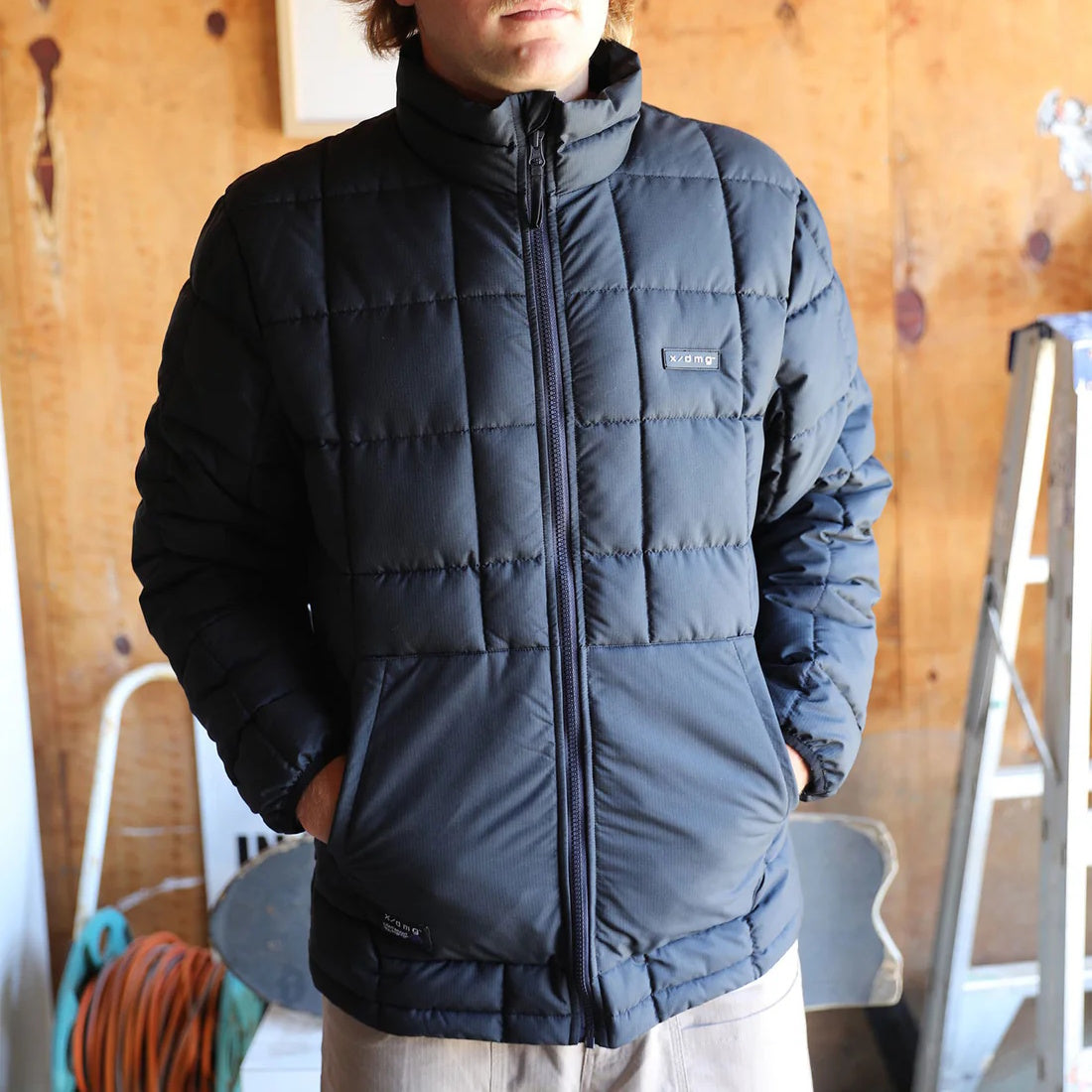 Person wearing a black X/DMG puffer jacket indoors with wooden walls in background.