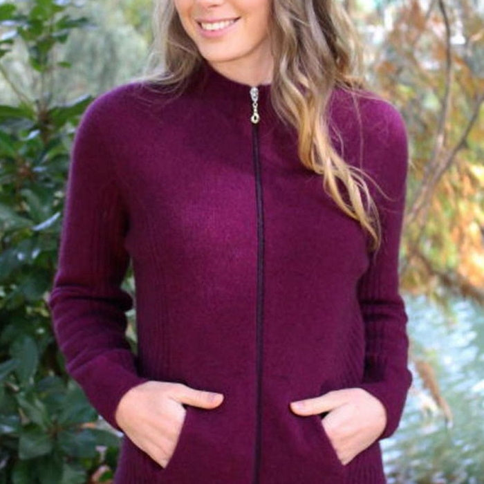 Female model wearing Lothlorian full zip possum merino jacket in Berry. The model has her hands in both pockets and the background is a riverbank.