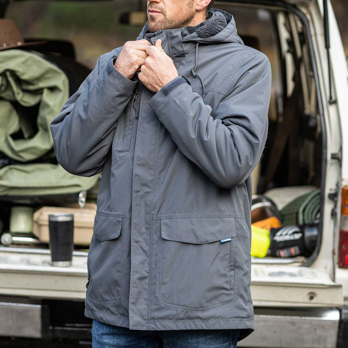 Male model wearing a Rainbird Altair Coat in Shadow. He is standing in front of a 4wd with boot open showing the camping goods inside.