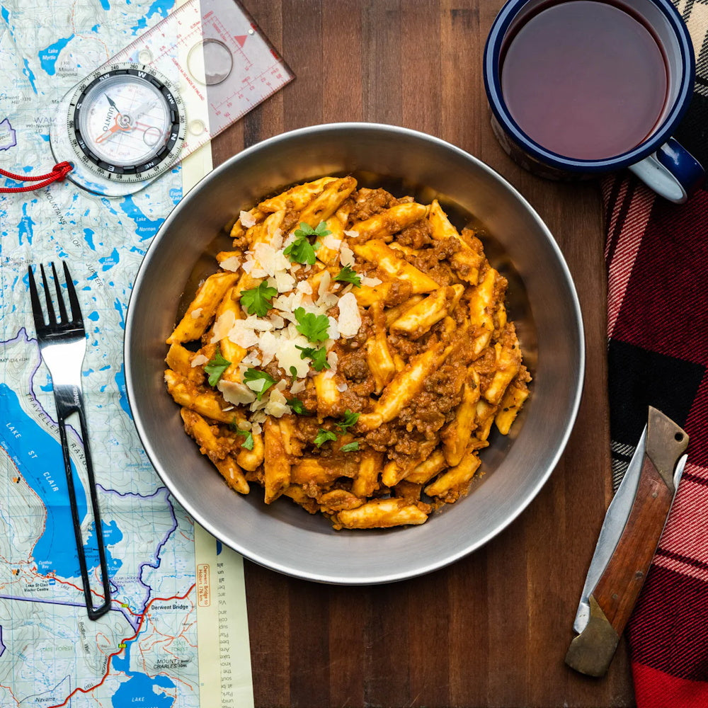 Bowl of Campers Pantry penne bolognese with a map, compass, and mug on a wooden board